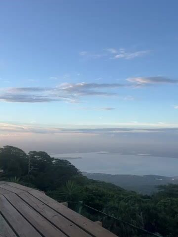 The video displays a scenic overlook with a wooden deck in the foreground. A vast body of water, possibly a lake or sea, stretches into the distance, dotted with islands. Lush green vegetation covers the surrounding hills and mountains. The sky above is a clear blue with scattered white clouds. Several warning signs are visible on a fence at the edge of the overlook. The footage captures a panoramic view of a natural landscape under a bright sky.