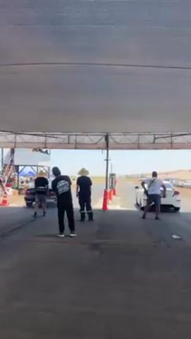 The video shows a white car accelerating down a drag racing strip. The car is seen from a low-angle perspective beneath a canopy. Four people stand to the left and right of the camera. Three of the people are wearing hats. The car speeds down the center of the track, passing orange cones on the left and right. A timing tower is seen in the background next to a short metal structure. People are visible in the background along the side of the track. The environment appears to be a rural area with a mostly dry landscape.