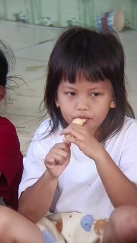 The video shows a young girl with dark hair and bangs, wearing a white t-shirt, eating a light-colored popsicle. She is holding the popsicle with both hands and bringing it to her mouth. Another child, wearing a red shirt, is partially visible on the left side of the frame. The background appears to be an outdoor or semi-outdoor setting with a tiled floor and a wall with a mural or painted design. Another young girl, wearing a maroon shirt, is also visible, with her eyes closed and holding what appears to be a piece of food.
