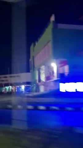 The video shows a nighttime view from a moving vehicle. The scene is mostly dark with artificial lights illuminating buildings and some traffic. The first building appears to be a modern structure with a green facade and a walkway. The next section shows a building under construction with exposed framework and lights. Then, a building with domes and arches is visible, along with some market stalls or shops. Finally, a prominent minaret and domes of a mosque or similar religious building are illuminated. The motion blur indicates the camera is in transit.