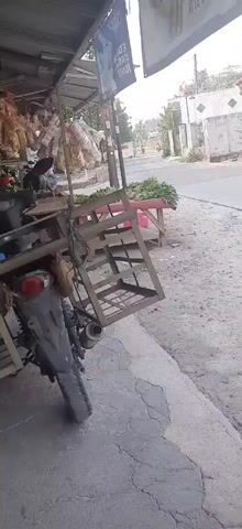 The video shows a street scene in what appears to be a rural or suburban area. On the left side of the frame, there is a roadside stall with a motorcycle parked next to it. The stall has various items displayed, including what looks like dried food products hanging overhead and vegetables on a table. A person is partially visible behind the stall. The motorcycle has a metal rack attached to its rear. A road runs through the middle of the scene. On the right side of the road, there are houses and a small shop with a sign that says "Yummy". A man is walking across the road, and a black car is parked in front of one of the houses. The ground is a mix of asphalt and dirt. The overall impression is of a typical street scene in a community with small businesses and residences.