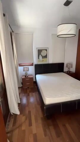 The video pans across a bedroom and into an adjacent room. In the bedroom, a bed with a white mattress and a black headboard is visible. There is a wooden nightstand with a lamp to the left of the bed, and a wooden wardrobe to the right. A ceiling light fixture with a decorative shade hangs above the bed. The floor is made of polished dark wood. The video then moves to a doorway, revealing a room with a black and white checkered floor. This room appears to be a kitchen or bathroom, with wooden cabinets and a window visible. An air conditioning unit is mounted on the wall above the doorway.