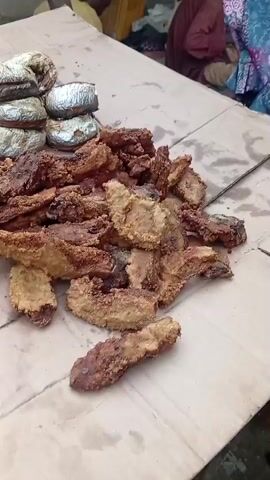 The video shows a close-up of a variety of fried food laid out on large pieces of cardboard on the ground. The food includes what appears to be fried plantains and possibly some other fried items, some of which are dark brown and crispy, while others are lighter in color. Several small, round, possibly baked or fried items are stacked in groups. A small clear plastic bag containing a light brown powder is visible on the cardboard. In some shots, a person's hands are seen near the food, but the person's face is not clearly visible. The background shows glimpses of what appears to be an outdoor market setting with other people and structures.