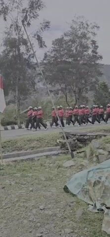 A group of people in matching red shirts and dark pants are marching in formation on a paved road. They are wearing white caps. A red and white flag is planted on a pole to the left of the frame. The road is lined with trees and vegetation, and a mountainous landscape is visible in the background. A motorcycle is parked on the left side of the road.