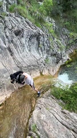 Two individuals are standing on a rocky ledge near a waterfall. The rock formations are intricately layered and textured. One person is wearing a white t-shirt, black shorts, a black cap, and red shoes. The other is wearing a black hooded jacket, black pants, and is using a walking stick for support. The person in the white shirt appears to be assisting the person with the walking stick, possibly helping them navigate the slippery rocks. A pool of clear water is visible at the bottom of the waterfall, surrounded by greenery. The environment appears to be a mountainous, natural setting with lush vegetation.