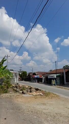 The video shows a street scene in what appears to be a village or small town. The sky is bright blue with scattered white clouds. Overhead, several power lines stretch across the frame, supported by utility poles. On either side of the paved road, there are buildings with tile roofs, some of which appear to be shops or small businesses. There are also some trees and vegetation visible. The overall impression is one of a sunny day in a rural or semi-rural setting.