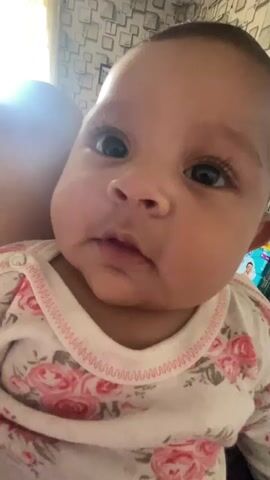 A close-up shot focuses on a baby looking at the camera. The baby has a light complexion, dark eyes, and full cheeks. They are wearing a white onesie with a pink floral pattern and a pink zigzag trim around the neckline. In the first few seconds, the baby's eyes are wide open, and they briefly smile, showing their tongue. Then, the baby's eyes widen, they look around, and their mouth opens slightly, revealing their tongue again before the video ends. A faint light is visible in the background on the left side, and a patterned wall can be seen behind the baby's head.