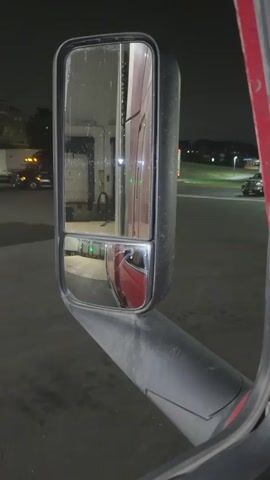The video shows a close-up view of a truck's side mirror at night. The mirror reflects a loading dock area with several trucks and trailers. The loading dock doors appear to be open, and red lights are visible, suggesting activity. The ground is wet, indicating rain or recent washing. The overall environment appears to be a commercial or industrial setting, possibly a distribution center or truck stop. The footage is steady and focused on the mirror, with no discernible movement of people or significant action.