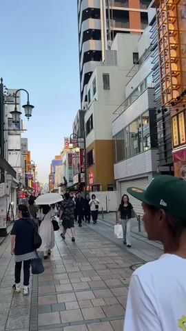 The video shows a busy street scene. The camera is looking from behind two people, one wearing a white t-shirt with a graphic and the other wearing a white t-shirt. People are walking along a paved sidewalk lined with shops. Some shops have awnings and display goods outside. There are signs in Japanese. The buildings are tall and modern. The sky is a pale blue. The camera pans slightly to the right. The time stamp is 00:00-00:05.