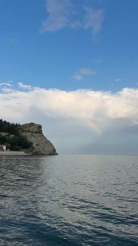 The video begins with a view of a calm sea under a bright blue sky with scattered clouds. A rocky cliff face with some vegetation and buildings is visible on the left side of the frame. As the video progresses, a faint rainbow appears in the sky behind the clouds. The camera pans slowly to the right, revealing more of the open sea. A person is visible swimming in the water in the distance towards the right side of the frame. The water is a deep blue-green with small ripples. The overall scene depicts a serene coastal landscape with a hint of a rainbow.