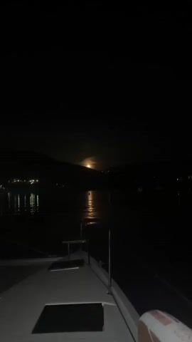 The video shows a serene night scene from the perspective of a boat on a body of water in Bigova, Montenegro. The foreground reveals the bow of a boat, with railings and a hatch visible. In the mid-ground, the dark water reflects the bright, full moon positioned just above a silhouette of hills or mountains in the background. A shimmering path of moonlight stretches across the water towards the boat. Distant lights from a settlement or other boats are faintly visible on the left side of the frame, also reflecting on the water. The camera slowly pans upwards, allowing for a better view of the moon and its luminous reflection, before panning back down slightly. The overall atmosphere is peaceful and calm.
