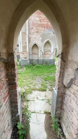 The video shows the interior of a ruined brick building, possibly a church, with arched doorways and windows. The walls are crumbling, exposing the red brick underneath, and some areas are covered in plaster that is also peeling away. The floor is paved with stones, but much of it is covered in grass and weeds. In the distance, through an opening in the wall, a small wooden structure with a gabled roof can be seen, along with trees and a cloudy sky. The overall impression is one of decay and abandonment.