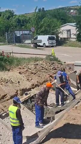 A construction crew is pouring concrete in a trench. There are five men working. One man in an orange hat is shoveling concrete. Three men in blue shirts are using shovels and other tools to spread the concrete. One man in a yellow vest is standing and watching. In the background, there is a white van, a white building, and a hill with trees. The sky is blue and it appears to be a sunny day.