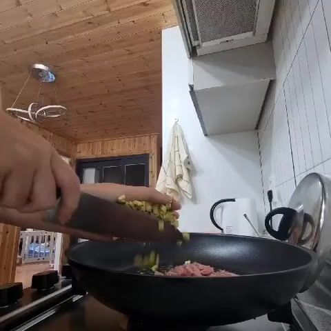 The video shows a close-up of a person's hands preparing food in a kitchen. The person is using a large knife to add chopped green vegetables to a frying pan that already contains pink meat. The kitchen has wooden paneling on the ceiling and walls, and white tiled backsplash. There is a range hood above the stove.