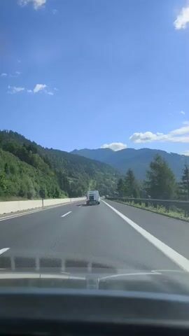 The video shows a view from inside a vehicle driving on a highway. The highway has multiple lanes and is bordered by a guardrail on the right and a concrete barrier on the left. Green forested mountains rise steeply on the left side of the highway, and more distant mountains are visible in the background. The sky is blue with scattered white clouds. A white camper van is visible ahead, traveling in the same direction in the right lane. A red car is also visible briefly in the left lane ahead of the camper van. The road appears to be wet in some areas. The timestamp indicates the video is from October 2022.