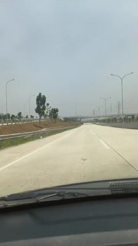 The video shows a first-person perspective from inside a vehicle driving on a highway. The highway is paved and has multiple lanes marked with white lines. There are streetlights and power lines visible along the side of the road. Vegetation, including trees and some construction or grading work, is present on the left side of the highway. A white car passes on the right side of the highway. The sky is overcast or hazy. There are no other people visible in the video. The scene appears to be during daylight.