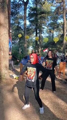 Two women in black shirts and leggings are dancing outdoors. The woman in the foreground wears a red hijab and white sneakers. The woman in the background has dark, curly hair and wears similar attire. Other people are visible in the background, some standing and some sitting on chairs. Tents and trees are present, and yellow lanterns are strung between the trees. The ground is dirt and covered with fallen leaves. The scene appears to be a casual outdoor gathering or event.