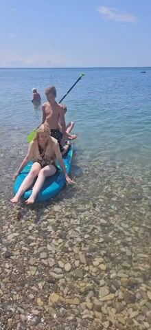 Two young people, a boy and a girl, are sitting on a paddleboard near the shore. The girl is in the front, and the boy is behind her. Another boy is kneeling behind them, holding a paddle. The paddleboard is in shallow, clear water with a rocky bottom. In the background, a person is standing in the water. The sky is clear and blue, indicating a sunny day. The scene appears to be a recreational activity at a beach or lake.