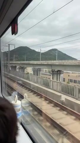 This video shows a view from inside a train traveling on a cloudy day. The camera focuses on the window, revealing railway tracks and a bridge structure outside. In the foreground, the back of a person's head and shoulders are visible, along with their hands holding a smartphone. Another person's head is partially visible in the lower left corner. The person with the phone appears to be interacting with it, possibly taking a picture or video of the scenery. The landscape outside consists of green hills and urban infrastructure.