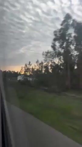 A vehicle is moving along a road, and the view out the window shows a rural landscape. There is a white building on a small hill surrounded by trees. The sky is filled with a textured, mackerel-like cloud pattern. A highway overpass is visible later in the clip.