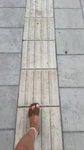 The video shows a sidewalk with tactile paving. There are people walking on the sidewalk, and a road with cars driving on it. There are also traffic signs and buildings in the background. The weather is overcast.