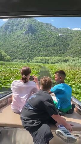 Three individuals are seated in a boat, facing away from the camera, looking out at a scenic landscape. The person in the center, wearing a black t-shirt, is leaning forward. The person on the left, wearing a pink shirt, is holding a phone up, possibly taking a picture or recording. The person on the right, wearing a blue shirt, is looking ahead. The boat is on a body of water covered with lily pads. In the background, a lush green mountain rises under a partly cloudy sky. The video appears to be a first-person perspective from someone in the boat, or an external shot of the boat moving through the water. The lighting suggests it is daytime.