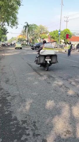 The video shows a street scene on a sunny day. A motorcycle is traveling down the road, carrying a large white package on the back. The motorcycle is in the foreground and is the main focus. In the background, there are other vehicles, including a car and several motorcycles. There are also buildings and trees lining the street. A traffic light is visible in the distance. The road surface appears to be asphalt with some wear and tear. The overall impression is of a typical urban or suburban street with moderate traffic.