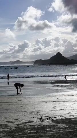 The video captures a serene beach scene at Selongbelanak, Indonesia, under a partly cloudy sky. The setting features a wide expanse of wet sand in the foreground, reflecting the bright sky. In the mid-ground, gentle waves lap at the shore, with the ocean stretching out to a horizon dotted with numerous small boats. Distant mountains rise prominently, forming a dramatic silhouette against the lighter sky.

Several individuals are present on the beach. In the immediate foreground, a person is initially seen bent down near the water's edge, then stands up and walks away towards the right. Further down the beach, a smaller figure, likely a child, is running playfully. To the left, two more people are visible in the shallow water near one of the boats.

The sky is filled with large, white and grey clouds, allowing patches of sunlight to break through and illuminate sections of the clouds and water. The overall lighting suggests it is daytime, possibly late afternoon, creating a soft, diffuse glow over the landscape. The atmosphere appears tranquil, with people enjoying leisure activities by the sea. No text is visible in the video.