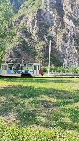 In Cheremushki, Russia, a tram moves along its tracks. The tram is multi-car and white with red accents. It appears to be decorated with various images on its side, some of which are obscured by the sun's glare. A few passengers are visible through the windows, though their features are indistinct.

The tram is set against a backdrop of a steep, rocky mountainside. A tall, lattice-style electricity pylon stands to the right of the tram, suggesting the presence of electrical infrastructure. Lush green grass covers the foreground, with some smaller trees and bushes interspersed. The lighting suggests it is daytime, likely morning or afternoon, with the sun casting shadows and creating bright highlights. The scene evokes a sense of peaceful transit through a scenic, natural environment, possibly a more rural or suburban area given the proximity of the tram line to the mountains. There is no visible text in the media.