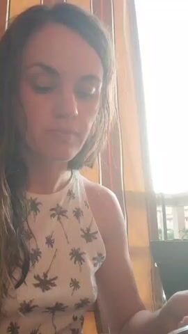 A woman with long dark hair is sitting at a table in front of a bowl of soup. She is wearing a white tank top with a palm tree pattern. She takes a spoonful of soup and brings it to her mouth, then eats it and chews. She is holding a spoon in her right hand. The background is blurred and appears to be a room with a window.