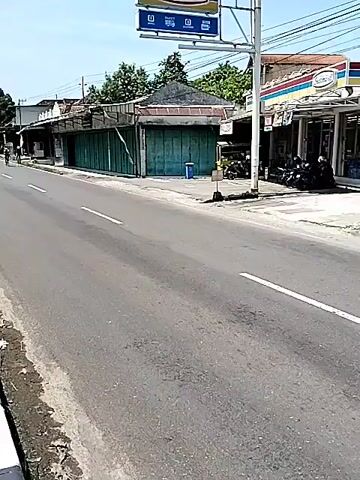 The scene is a road in Indonesia, in daylight and under a bright sky. Three cyclists are riding towards the camera on the right side of the road, along the solid white line separating the lane from the other. One cyclist is slightly ahead of the other two. A scooter passes them on the left side of the road. The cyclists appear to be wearing helmets and cycling gear.

On the right side of the road is an Indomaret convenience store with a sign visible at the front. Adjacent to it is a building with closed green roll-up doors, seemingly a shuttered shop. A small trash can sits in front of this building. Next to the Indomaret store is a green and white entrance structure. Parked motorcycles are visible near the store. The street appears to be well-maintained with painted lane markings. The area has scattered buildings, trees, and telephone poles.