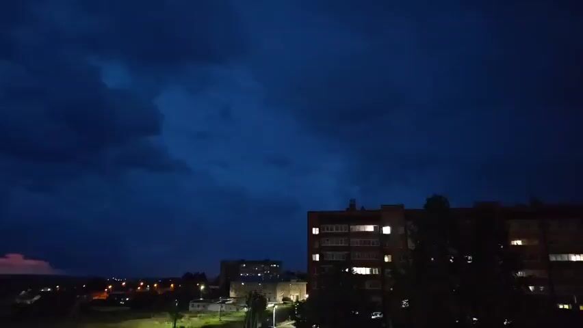 A nighttime scene depicts a dark, cloudy sky with a distant cityscape below. A tall apartment building with illuminated windows is prominently visible on the right side of the frame, with trees in the foreground. In the distance, other buildings are visible, along with streetlights. A faint pinkish glow can be seen on the horizon to the far left, possibly indicating a distant event or light source. The sky is a deep blue, suggesting twilight or a stormy evening. The overall atmosphere is dark and somewhat ominous.