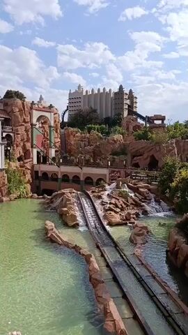 The video shows a water ride at a theme park with a log flume. The flume goes through a rocky landscape with waterfalls and lush greenery. In the background, a large, multi-story building with a distinctive architectural style resembles a palace or temple. The sky is blue with scattered white clouds. The water in the flume is a light green color. The ride appears to be empty at this moment.
