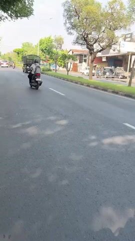 A scooter rider travels down a paved road. The rider is wearing a dark jacket and a helmet. A large green truck is in front of the scooter. The road is lined with trees and buildings. The sky is partly cloudy.