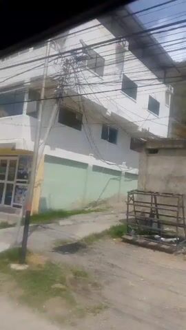 The video pans across a street scene. The first shot shows a multi-story building with shops on the ground floor. The second shot shows another building with barred windows and shuttered storefronts. The third shot shows a dirt road winding through lush green vegetation and a stone wall, with a glimpse of a hill in the background. The fourth shot shows more vegetation and a hill. The final shot shows a street with a car parked on the left, a concrete wall, a pole with wires, and more vegetation leading up a hill. The video appears to be filmed from a moving vehicle.