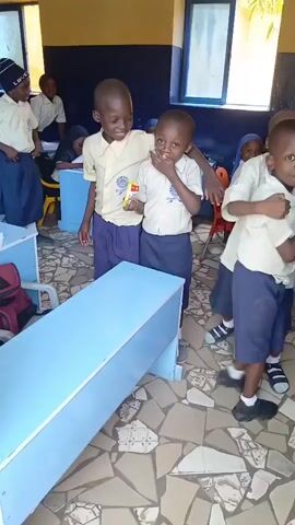 In a classroom, a group of about eight young children, wearing school uniforms consisting of light-colored shirts and dark-colored skirts or shorts, are present. Four boys stand close to each other in the foreground, with one having his arm around another. A fifth child appears behind the group. In the background, other children sit at desks. The classroom is furnished with light blue desks. The floor is covered with a mosaic tile pattern. One child is holding a snack packet. There is natural light from the windows in the background. The children are looking towards the camera, smiling and interacting with each other.