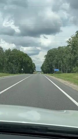 The video shows a point-of-view shot from inside a vehicle traveling on a paved road. The road is flanked by dense green trees on both sides, creating a tunnel effect. The sky above is overcast with large, dark clouds. There are white lane markings on the road. In the distance, there are faint signs and what appear to be a few other vehicles on the road. A white sign with blue text is visible on the right side of the road. The front of the vehicle, including the dashboard and windshield, is visible in the foreground. The overall environment appears to be rural or suburban.