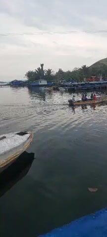 A small aluminum boat with an outboard motor is moving across calm water. The boat carries four people, two adults and two children. The boat is heading away from the camera towards a shoreline with structures and a hill in the background. The water is dark and reflects the overcast sky and the surrounding environment. The shoreline appears to be a small village or fishing settlement with wooden piers and buildings. The sky is cloudy, suggesting either early morning or late afternoon.