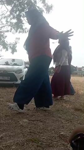 A group of people are outdoors near a car. The person in the foreground on the left is wearing a red shirt and dark pants. They appear to be dancing or exercising. Another person in the middle ground is wearing a white top and a long maroon skirt, also in motion. A third person is partially visible behind them, wearing a dark headscarf. The ground is covered with dry grass and leaves. A white car is parked in the background. There are trees and vegetation surrounding the area, with a bright, overcast sky. A red and white checkered blanket and a bottle of water are visible in the lower left corner of the frame.