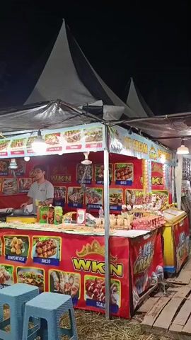 A nighttime shot of a food stall at an outdoor event. The stall is decorated with red banners featuring images of various food items like skewers, meatballs, and ice cream. A young man wearing a grey shirt stands behind the counter, preparing food. Several blue stools are placed in front of the stall. Tents and lights are visible in the background, suggesting a festive atmosphere. The video captures the food stall in operation, with the vendor attending to customers.