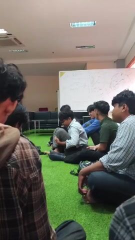 The video shows a group of young men sitting on a green carpeted floor in a room. A whiteboard covered in writing is visible in the background, along with some furniture and ceiling lights. Some of the men are looking at each other, and one appears to be talking. The camera is focused on the group from behind, with a person in the foreground wearing a plaid shirt and hoodie. The overall atmosphere appears casual and relaxed.