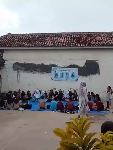 The video shows a group of children sitting on a blue tarp in an outdoor space. They appear to be attending an event or gathering, possibly at a school or community center. A woman in a white hijab stands among them. A banner is attached to the building behind the children, which appears to have some images and text about sanitation. Some children are walking around the seated group. The location appears to be in a residential area, with a building with a red tile roof and landscaping. The weather looks overcast.