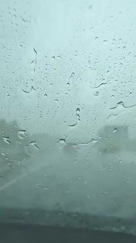 This is a video captured from inside a vehicle driving on a highway during a rainstorm. The primary subject is the car windshield, covered in numerous raindrops of varying sizes, distorting the view of the outside world. The scene outside is hazy and obscured by the rain, with the faint shapes of other vehicles and the road visible. The weather is clearly inclement, with heavy rain creating a gray and muted atmosphere. The overall mood conveyed is one of driving through challenging weather conditions, potentially with a sense of introspection or focus on the road ahead. The time of day is unclear due to the overcast sky. There are no discernible people or specific objects within the frame other than the raindrops and the interior of the car in the foreground. No visible text is present in the video.