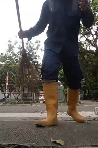 A person wearing a hard hat, mask, and yellow boots is sweeping debris off a paved surface with a broom. The person is dressed in dark clothing. The background shows trees, a building, and a red and white barrier.