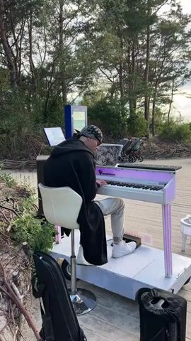 A man wearing a black hoodie, black cap, and white sneakers plays a portable electronic keyboard with purple lighting. The keyboard is mounted on a wheeled platform, and the man is seated on a white stool. He is wearing light-colored pants and has a black cloth draped over his shoulders. The keyboard has a laptop placed on top of it. The scene is outdoors, on a wooden boardwalk, with a sandy area and trees to the left. In the background, several people are walking, some with strollers, and others are sitting at tables. A child is riding a scooter towards the camera. The sky is cloudy with sunlight breaking through, and a pier extends into the water in the distance.