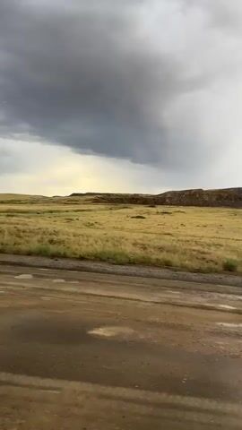 A dirt road, wet from recent rain, stretches across the foreground of the image. Beyond the road, a vast, flat field of dry, golden grass extends to a low, rugged ridge in the mid-ground. The sky above is dramatic and overcast, with heavy, dark storm clouds dominating the left side and lighter, brighter clouds on the right, where sunlight is breaking through. The overall scene depicts a rural, open landscape under a changing sky. The weather appears to be a transition from or towards a storm. The time of day is ambiguous due to the cloud cover, but the brighter patches suggest it could be late afternoon or early evening. There is no visible text in the image.
