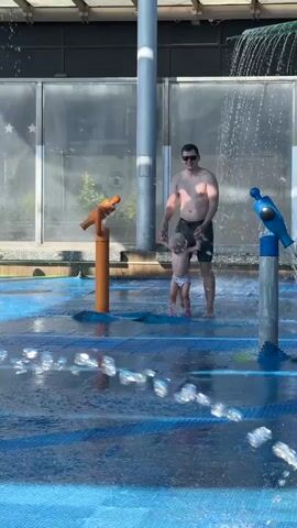 A father and a young child are standing in a splash pad. The child is wearing a swimsuit and holding the father's hand. The father is shirtless and wearing swim trunks. They are surrounded by water features, including spray jets and a waterfall. The splash pad appears to be made of blue rubber material. In the background, there is a building with large glass windows. Another child can be seen walking in the background.