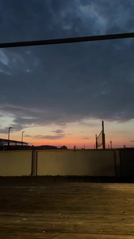 The media file captures a tranquil sunset scene, likely at a beach or outdoor recreational area in Singen (Hohentwiel), Germany. The sky is a canvas of dramatic colors, transitioning from deep blues and grays at the top to vibrant oranges and pinks near the horizon, indicating the time of day is dusk. Scattered clouds add texture to the atmospheric display.

In the foreground, a wooden deck provides a vantage point for viewing the scene. A light-colored railing separates the deck from the area beyond. Silhouetted against the colorful sky are various structures. To the left, what appears to be part of a building or sports facility with prominent light poles is visible. Further to the right, a volleyball net and its poles stand as a testament to the area's recreational purpose. A solitary figure can be faintly seen near the horizon, adding a sense of human presence to the expansive landscape.

The overall mood is serene and peaceful, evoking a sense of calm as the day draws to a close. The combination of natural beauty in the sky and the subtle human-made elements suggests a harmonious blend of nature and leisure. No visible text is present in the media file.