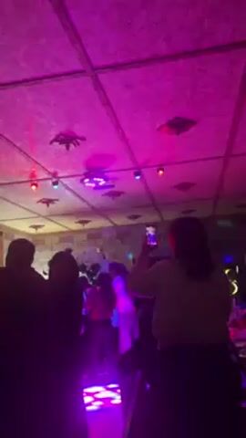 The video shows a lively party scene, with people dancing and socializing in a room lit by colorful disco lights. A person in the foreground holds up their phone, apparently recording the event. The ceiling is decorated with square panels, and additional light sources are visible, creating a vibrant and energetic atmosphere. The individuals seem to be enjoying themselves amidst the music and colorful lighting.
