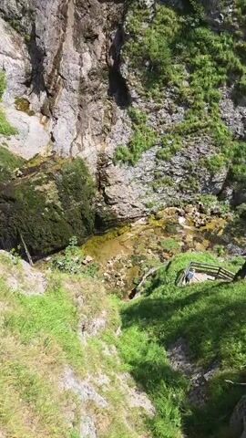 The video shows a waterfall cascading down a rocky, moss-covered cliff face surrounded by lush green vegetation. The perspective appears to be from a higher vantage point looking down at the waterfall and the surrounding natural landscape. The waterfall has multiple tiers and flows over rocks of varying sizes. The cliff is made of rough, grey rock with patches of green moss and grass growing on it. Trees and bushes are visible on the upper parts of the cliff and the surrounding terrain. The water at the base of the waterfall collects in a shallow, rocky pool. There are some wooden structures visible on the right side of the frame, possibly part of a viewing platform or walkway. The sunlight suggests it is daytime, likely a clear or partly cloudy day. No people are clearly visible in the frame, though there is a hint of what might be a person or two near the top of the waterfall on the left side.